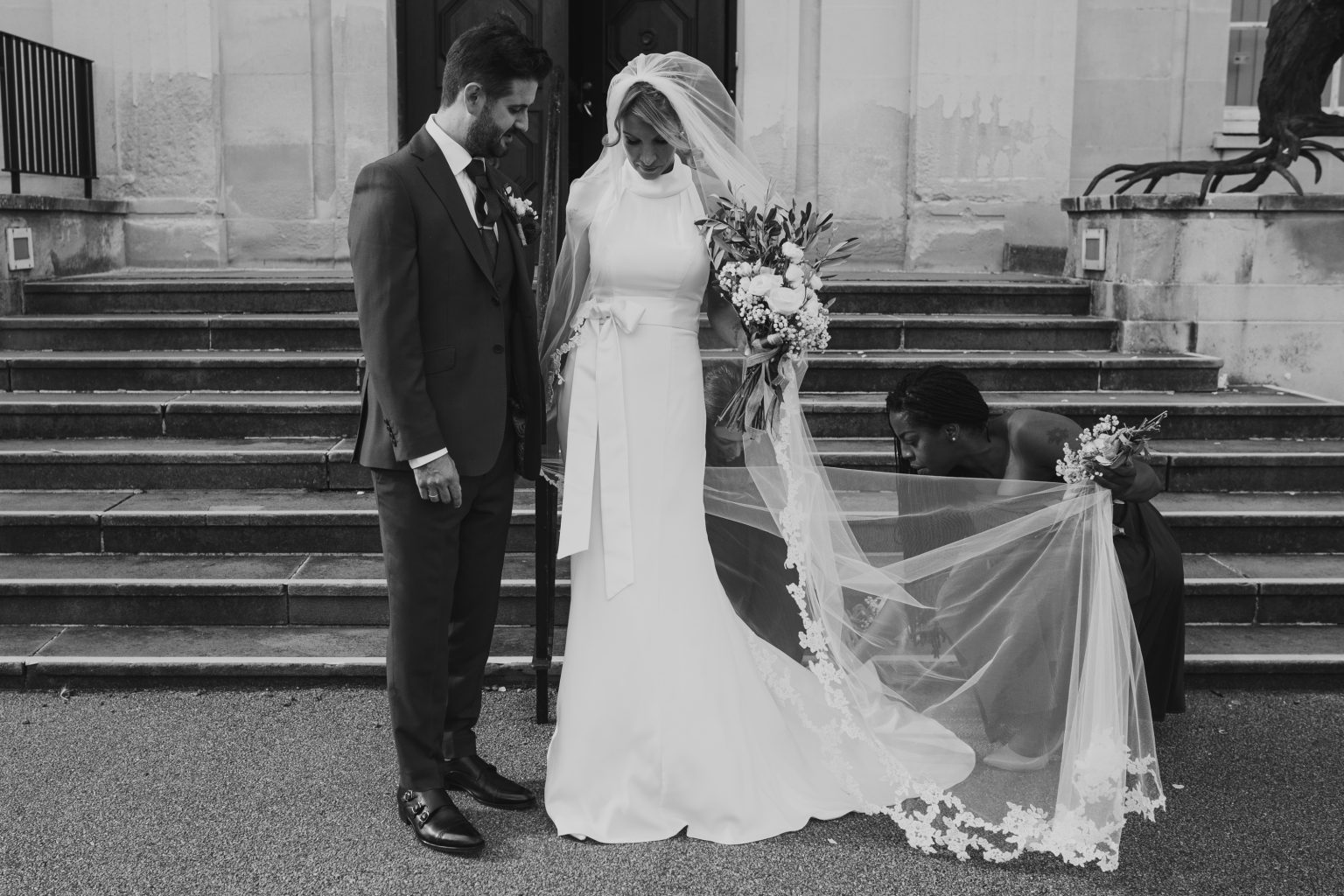 Bride and groom at the bottom of the steps of Hertford Council Chambers with bridesmaid fanning out the bride's veil, London wedding photography, Romantic London wedding, beautiful London wedding venue, Bride and groom London, London wedding photography inspiration, Elegant London wedding photos, Stylish London wedding portraits, London wedding photography packages, Chic London wedding photography, London wedding photography services London wedding photographer capturing love in the city, Elegant and romantic wedding photography in London, Beautiful bridal portraits in iconic London locations, London wedding photographer creating timeless memories, Stylish and sophisticated wedding photography in London, Breathtaking images of love and celebration in London, Award-winning wedding photography in the heart of London, London's top wedding photographer capturing your special day, Chic and modern wedding photography in London, Unforgettable moments captured by a London wedding photographer