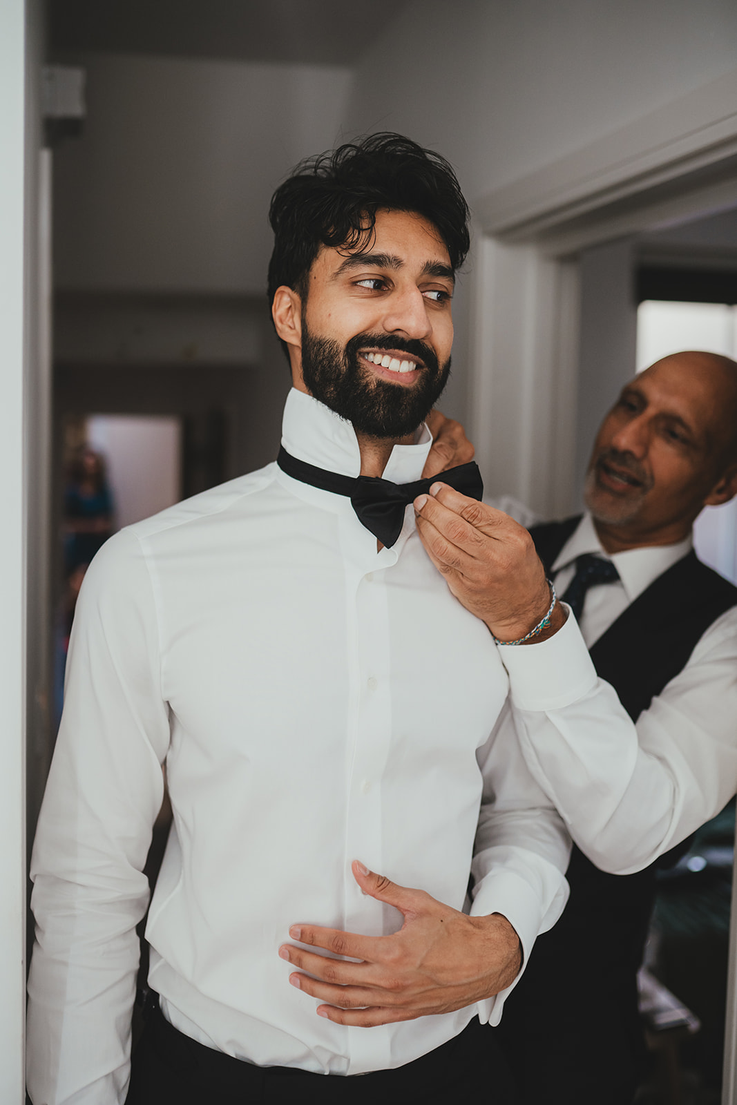 Groom smiling as someone adjusts his bow tie during getting ready.