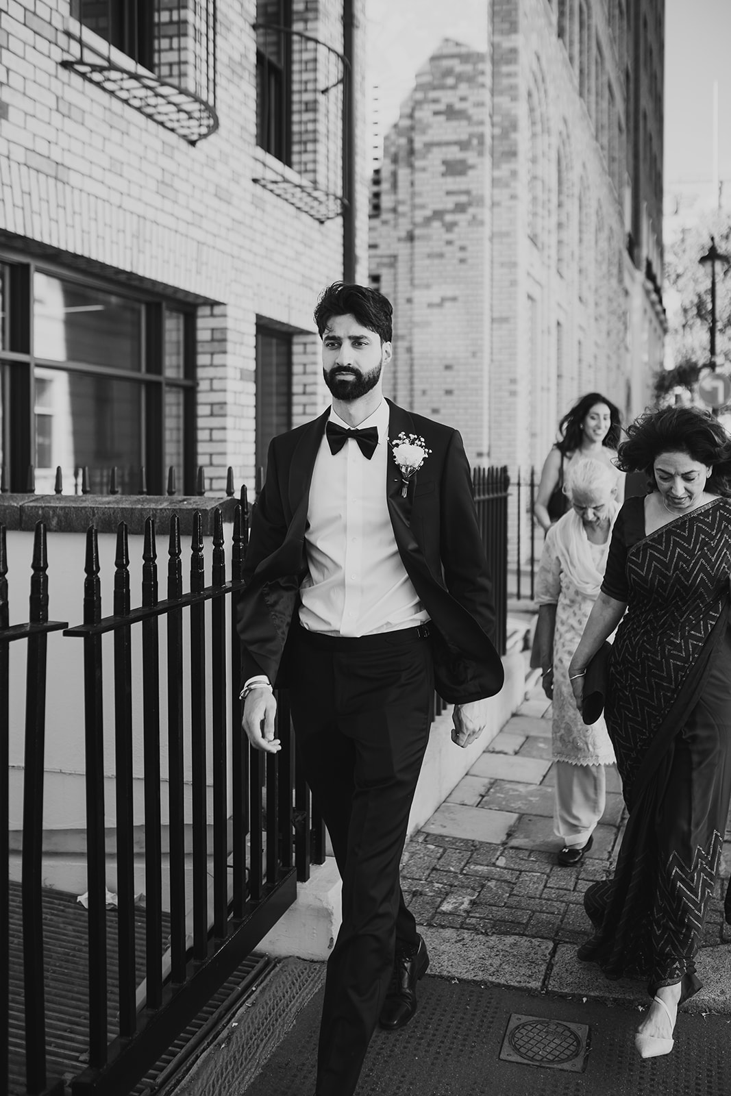 Groom in a tuxedo walking along a London street with family behind him.