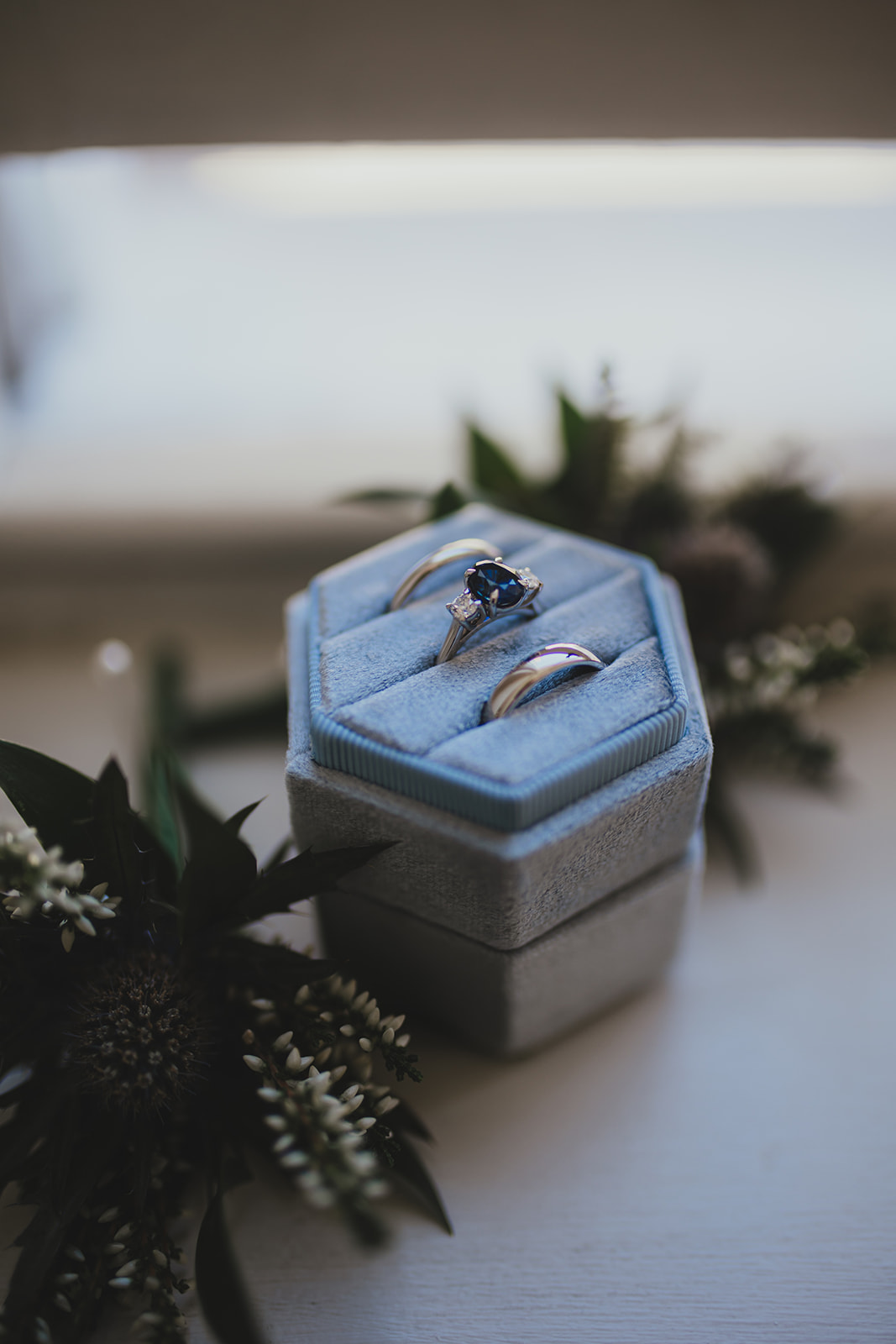 Wedding rings in a blue velvet ring box on a table with greenery.