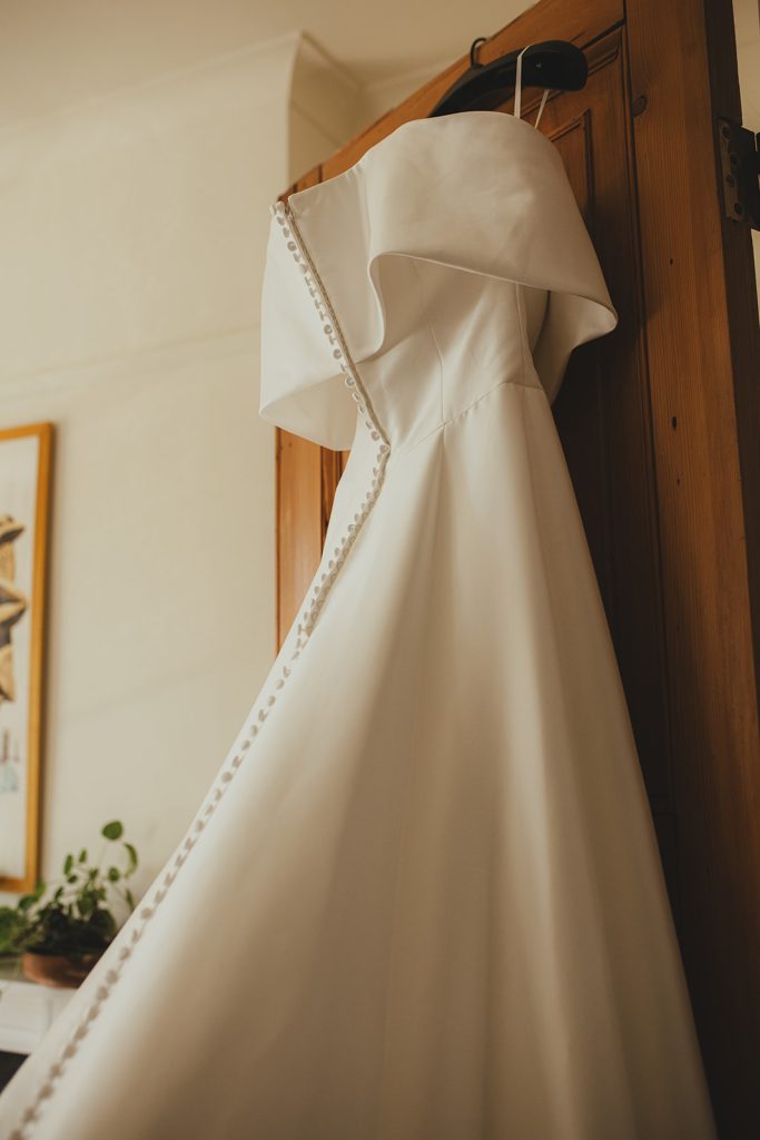 Off-the-shoulder wedding dress hanging on a wooden wardrobe in soft window light.