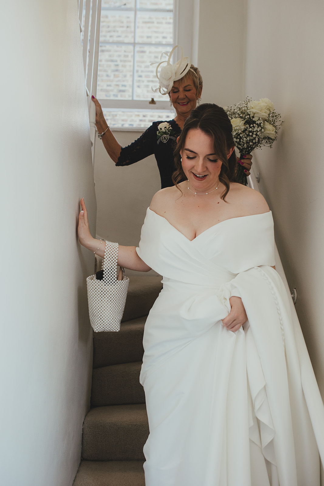 Bride walking down the stairs with a family member behind her holding flowers.