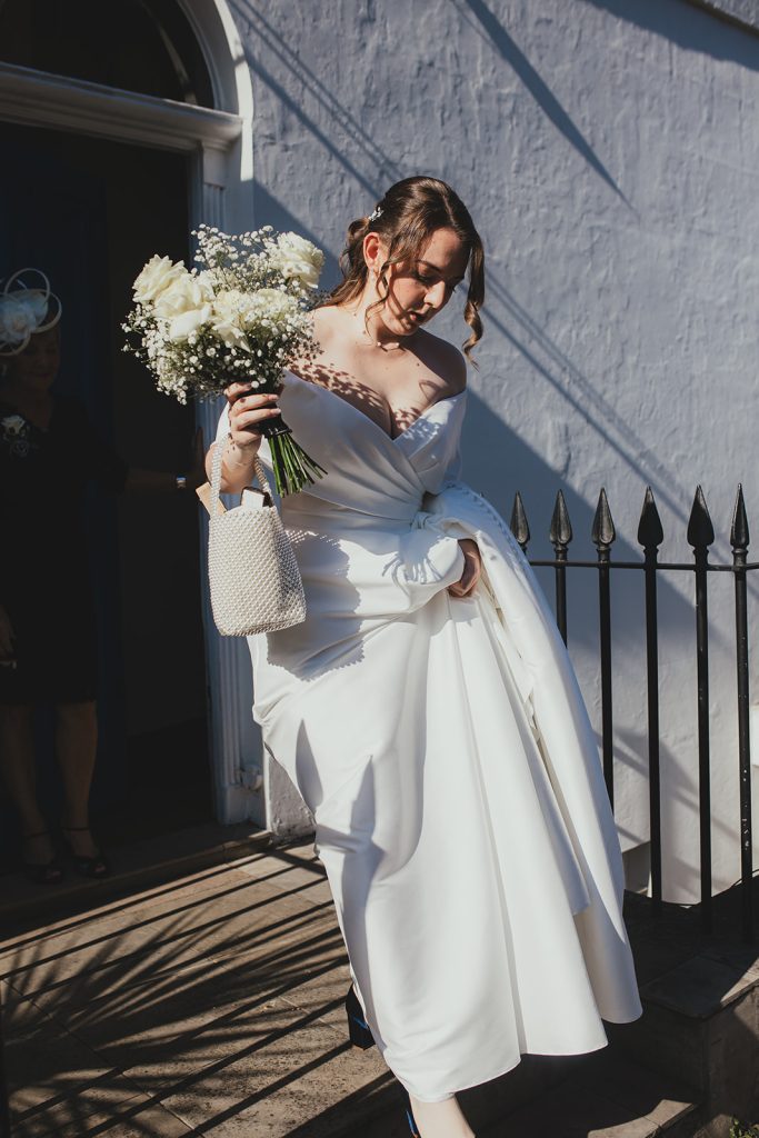 Bride stepping out into sunlight holding her bouquet outside the house.