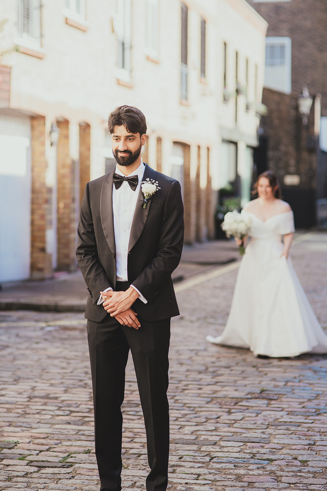 Groom waiting on a cobbled London street as the bride approaches from behind.