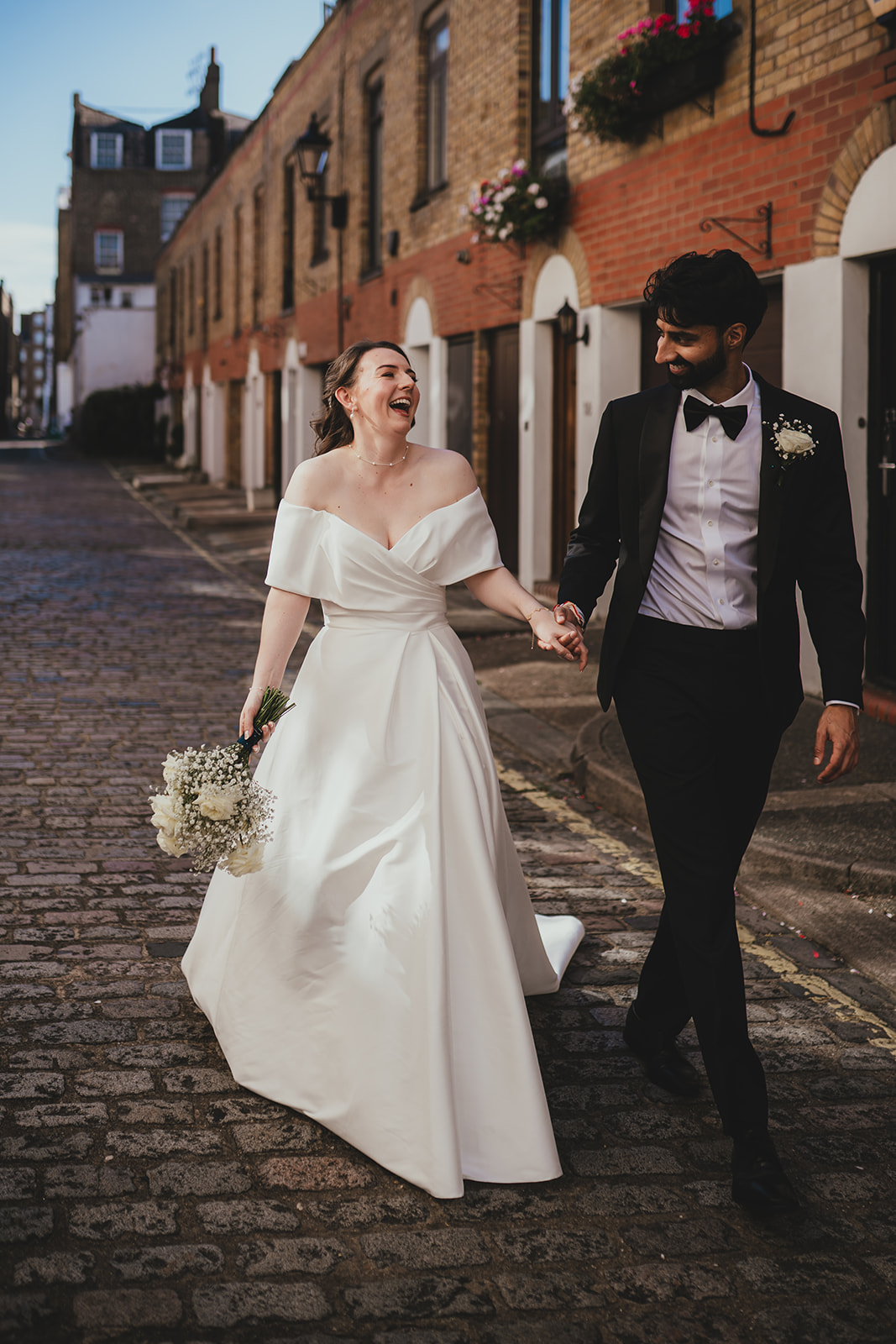 Bride and groom walk hand-in-hand along a cobbled mews street in London, smiling and laughing.