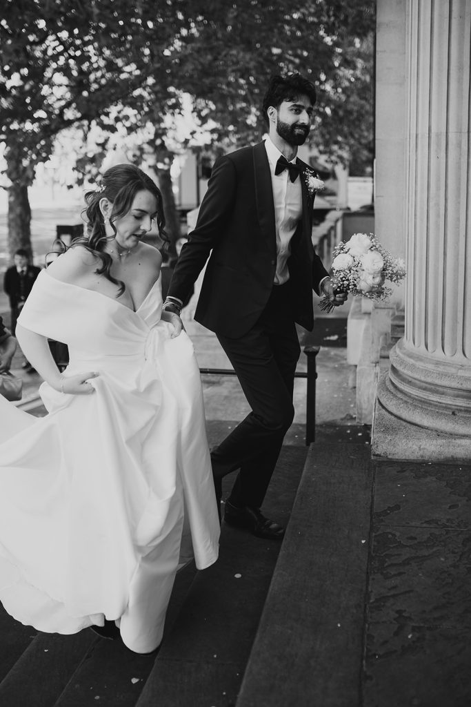 Black and white photo of the bride lifting her dress as she walks up the steps outside Old Marylebone Town Hall
