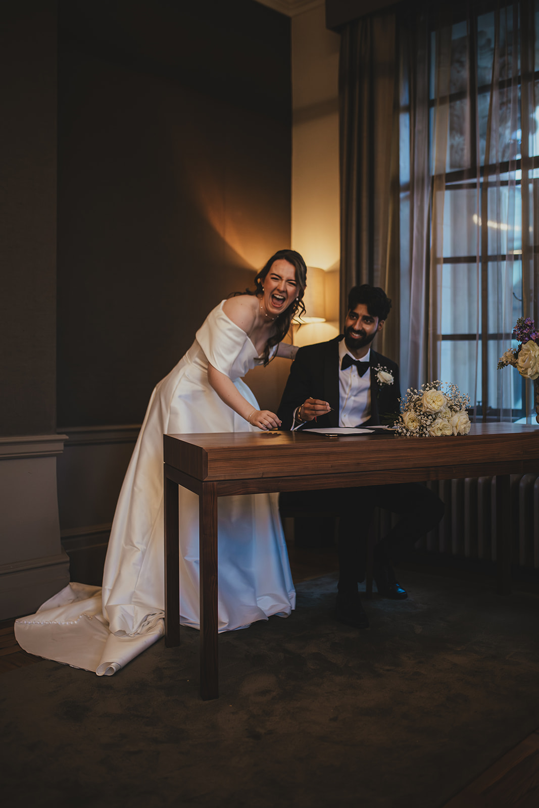 Bride laughs beside the signing table as the groom signs the register at Old Marylebone Town Hall