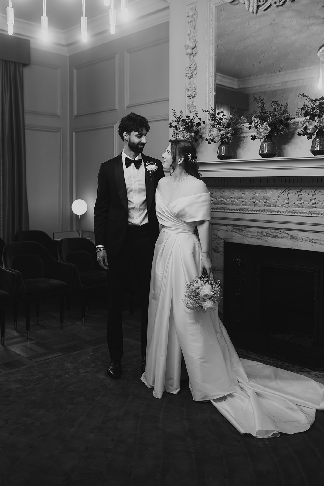 Black and white photo of the couple waiting in front of a flower-decorated mantelpiece at Old Marylebone Town Hall