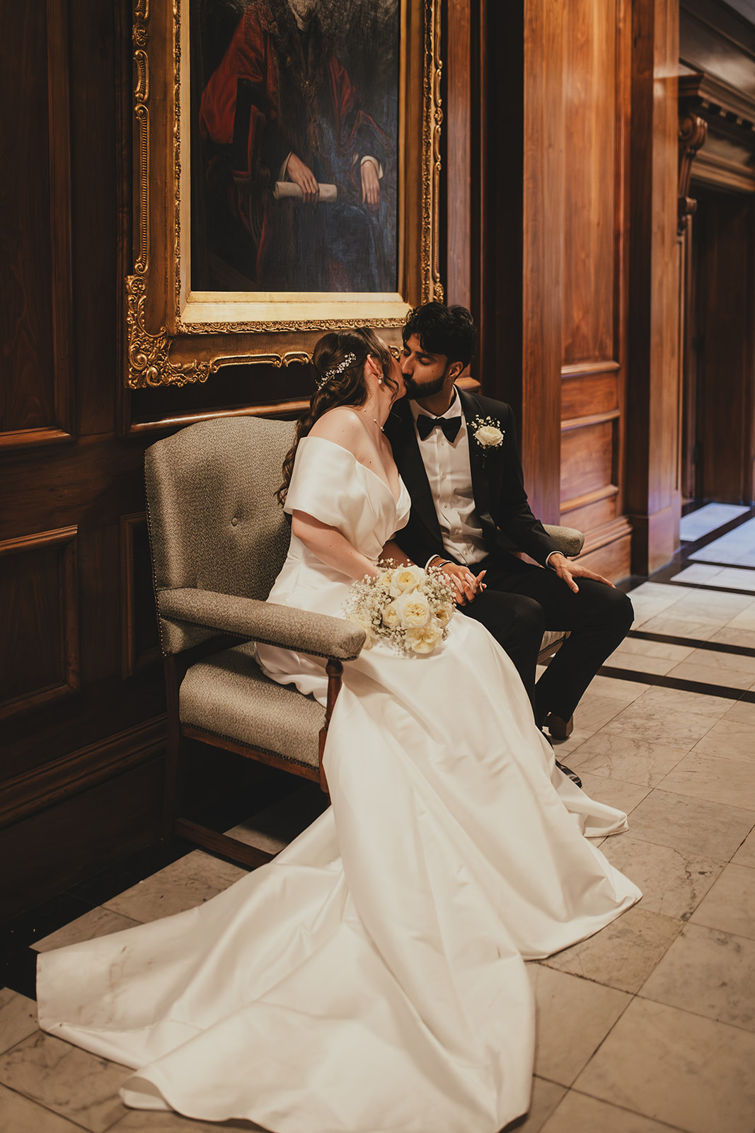 Bride and groom share a kiss on a bench inside Old Marylebone Town Hall, bouquet in the bride’s lap