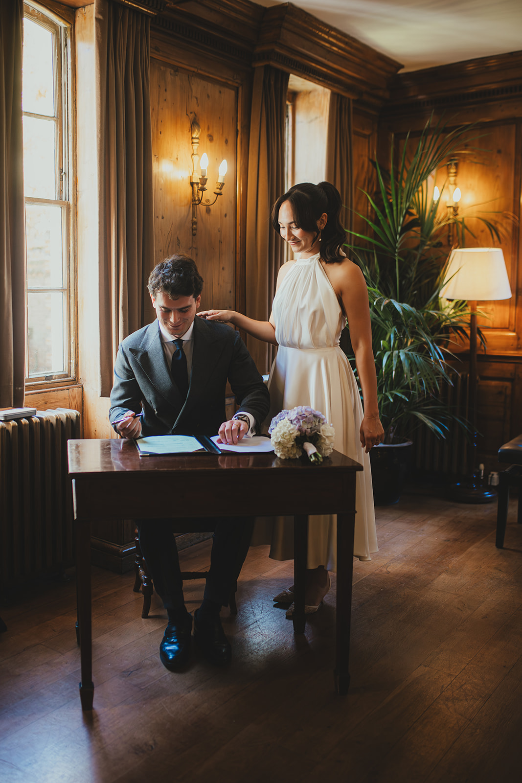 Bride standing next to grppm as he signs the wedding certificate at Brugh House 